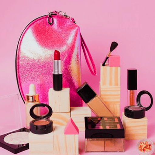 Bolsinha rosa metálica com maquiagem e produtos de beleza ao redor, fundo rosa pastel.