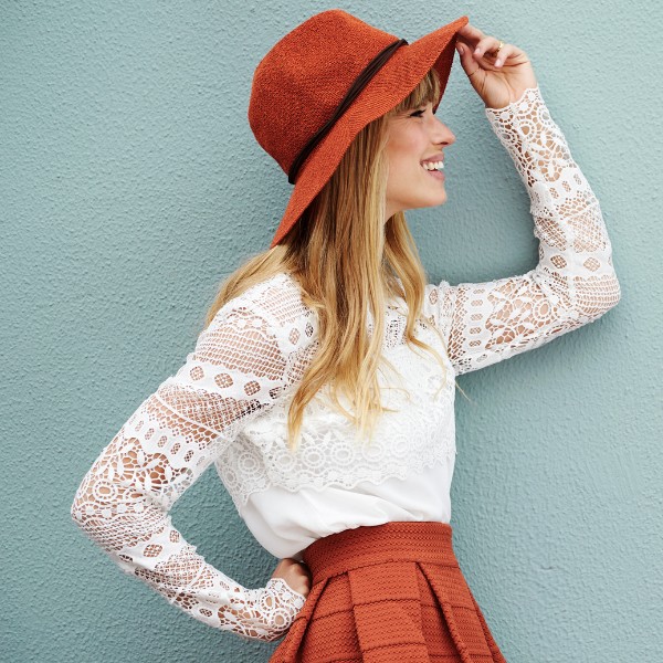 Chapéu de feltro laranja, mulher sorridente com blusa de renda branca e saia também laranja, estilo afetivo, fundo azul texturizado.